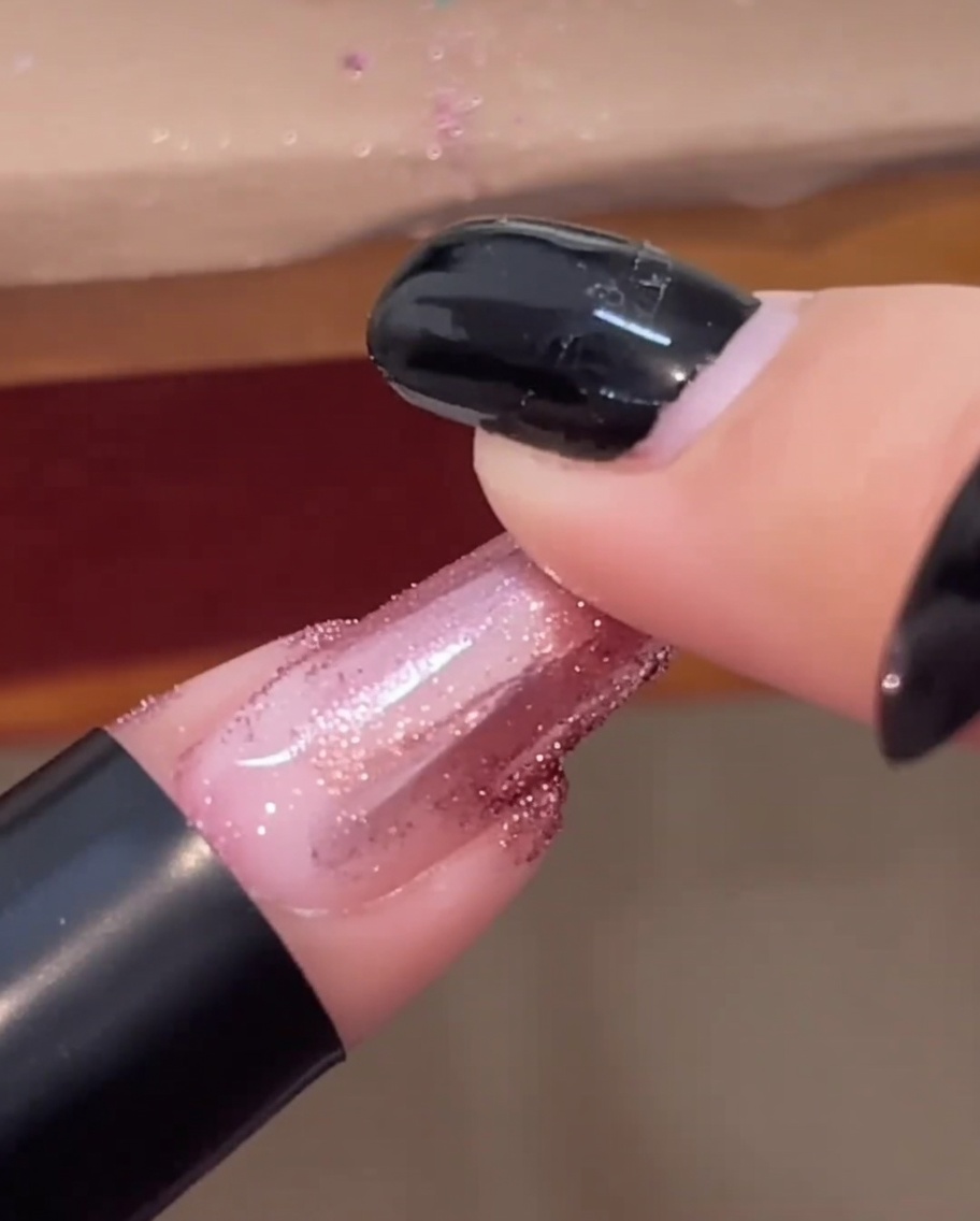 A fingertip rubbing rose gold chrome powder over a finished, glossy nail.