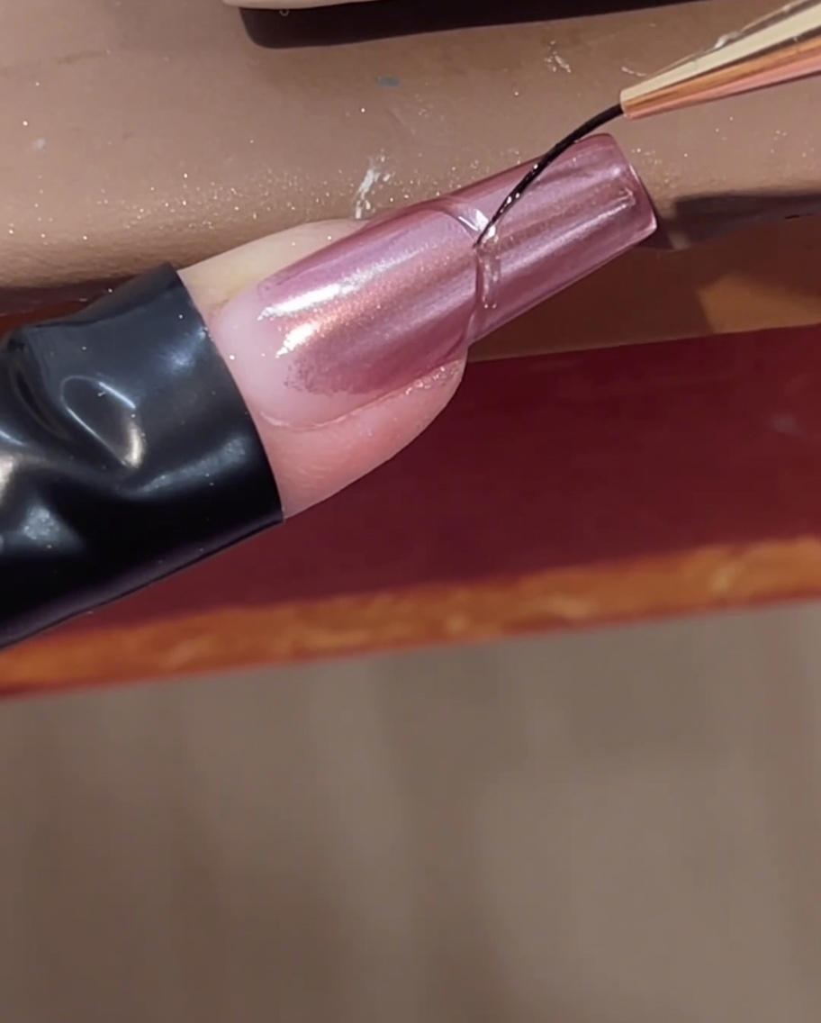 A fine liner brush adding a curved clear builder gel line near the cuticle of a pink chrome nail.