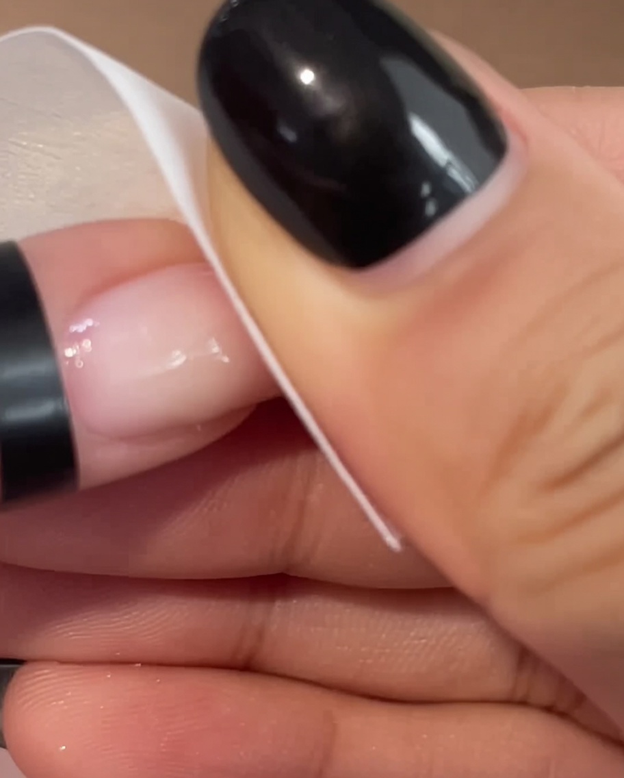 A close-up view of a hand using a white wipe to gently clean the surface of a pink manicured fingernail.