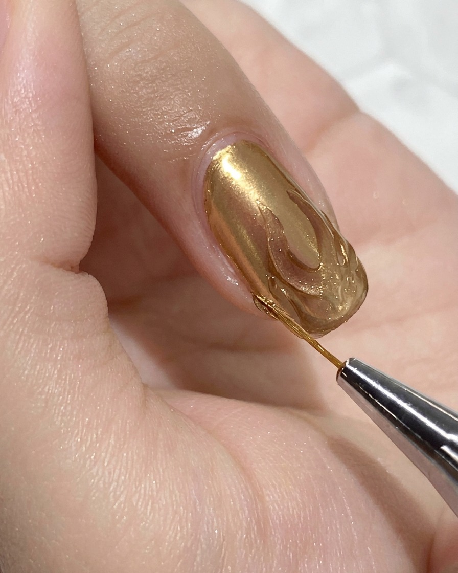 A close-up view of a fine detail brush applying clear hard gel top coat inside a gold chrome flame outline on a sheer pink fingernail.