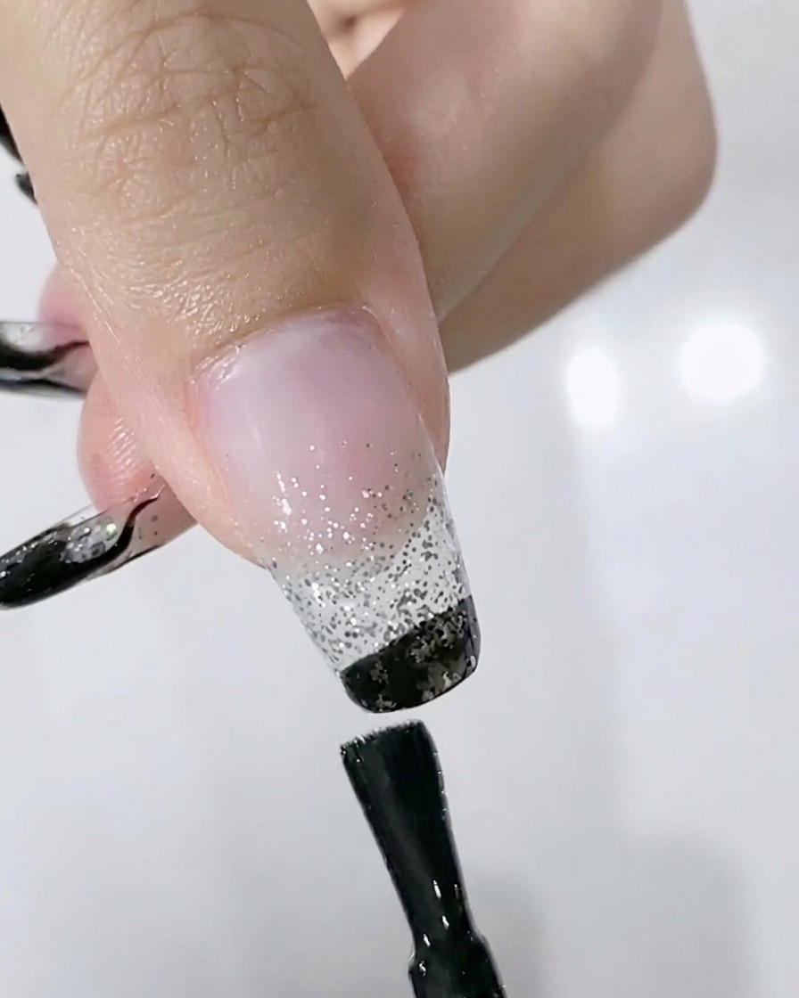 A brush applying black gel polish to the free edge of a nail over a silver glitter gradient.