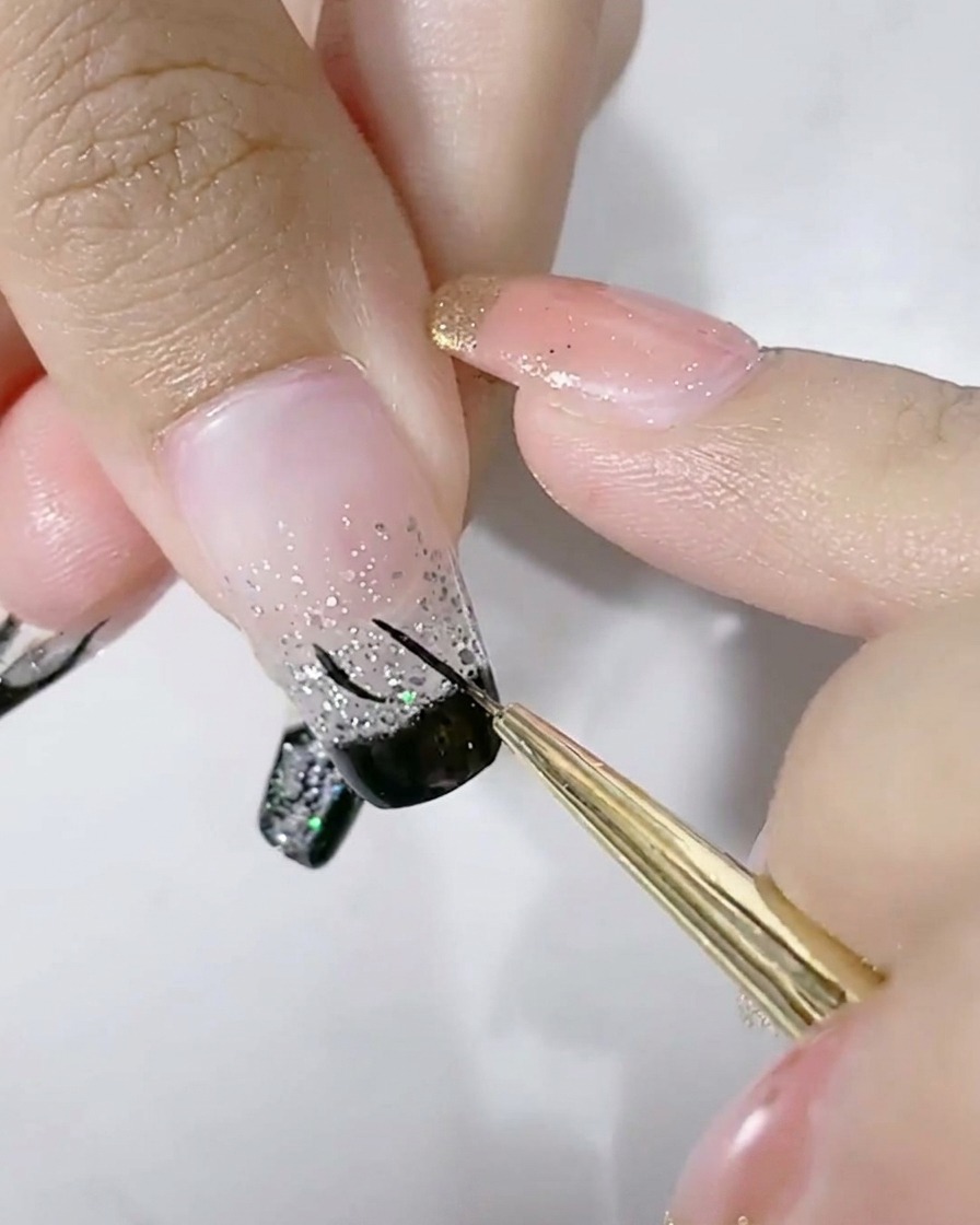 A fine liner brush drawing a black U-shape outline on the silver glitter portion of a fingernail.