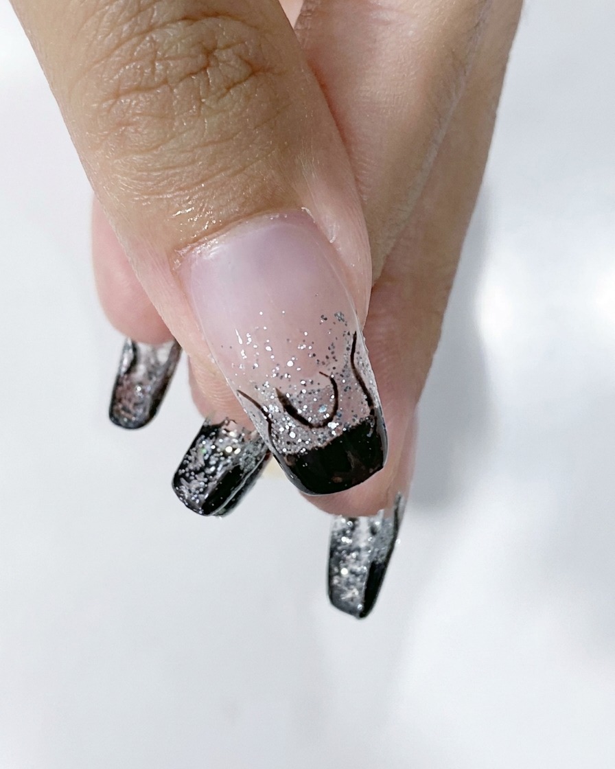 A close-up of a fingernail showing a completed black flame outline over a silver glitter gradient base.