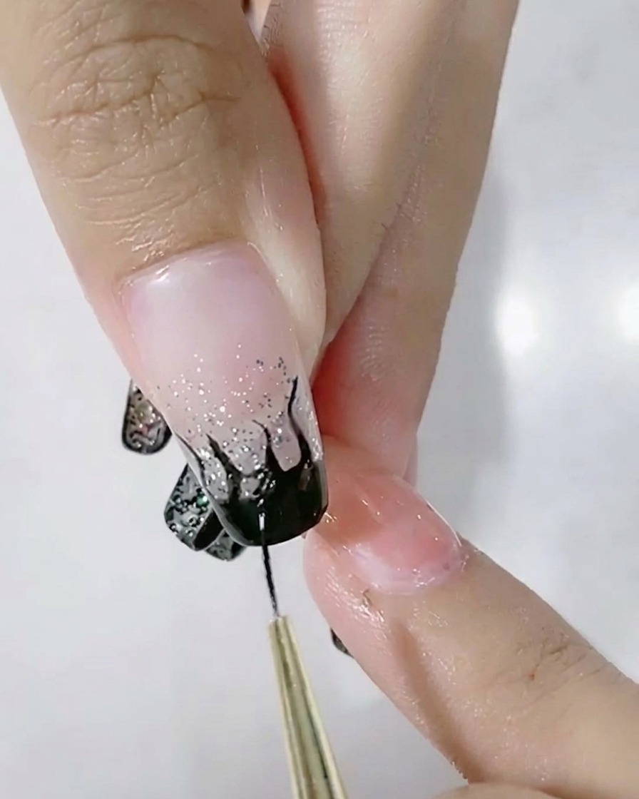 A liner brush filling in a black flame design on a glitter-gradient nail.