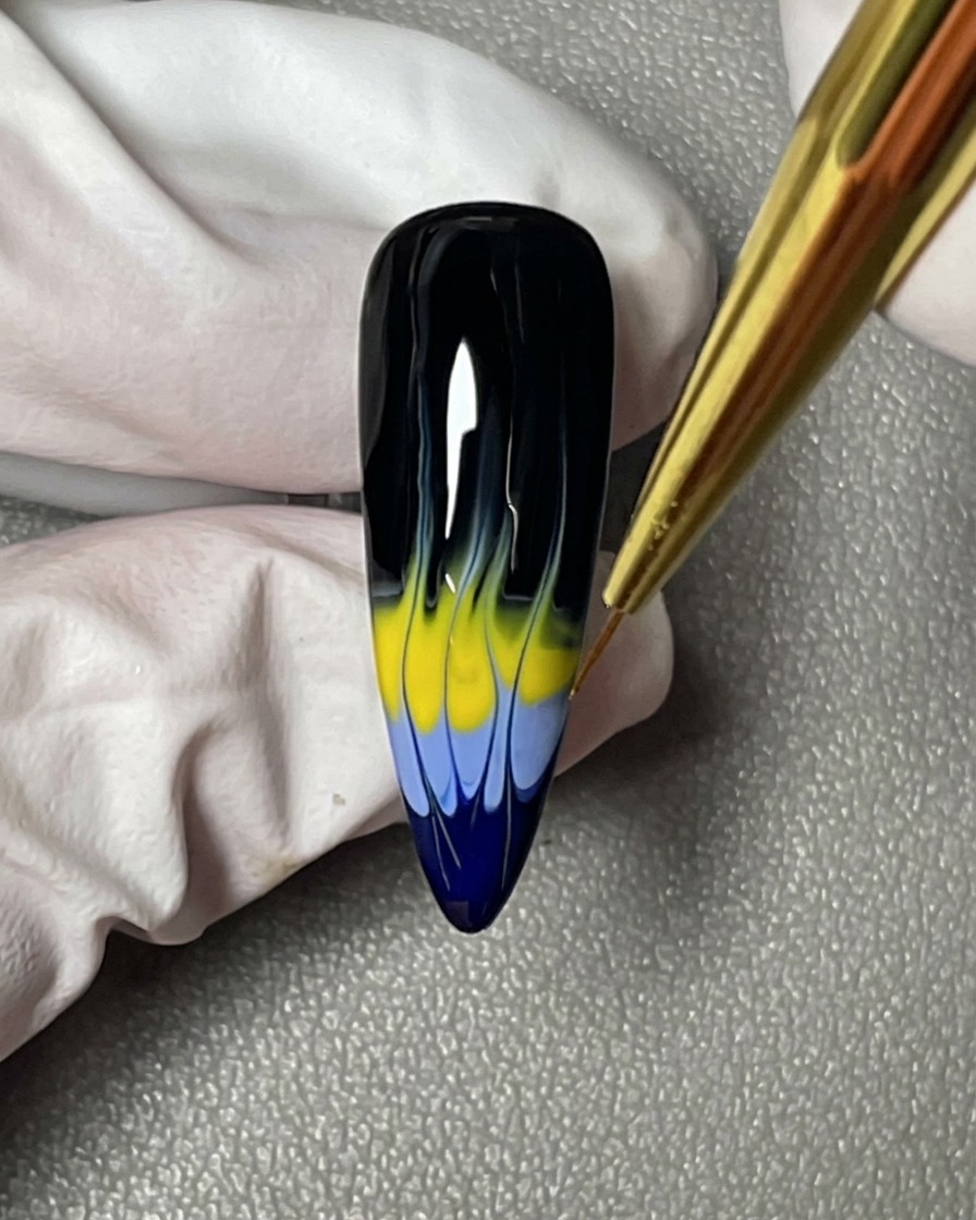 A close-up showing a liner brush perfecting the sharp, tapered points of the blue and yellow flame design on a black nail.