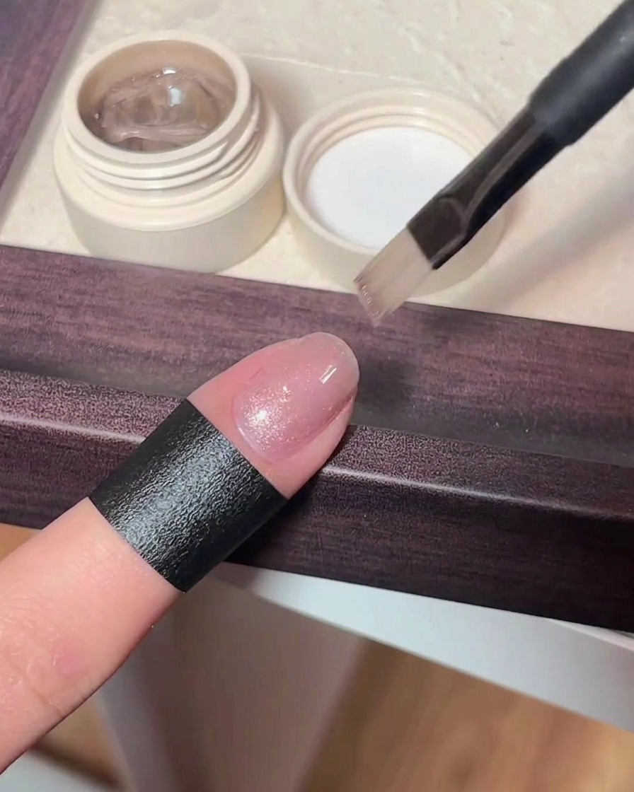 A close-up view of a brush applying a sheer, shimmering pinkish cat eye gel polish onto a natural fingernail.