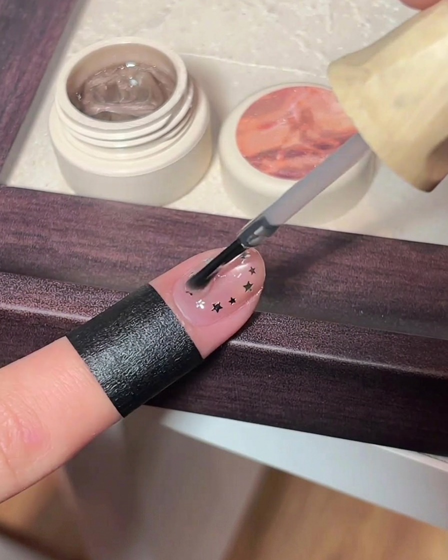 A nail brush applying a thick layer of clear top coat over a pink cat eye nail decorated with tiny metallic star decals.