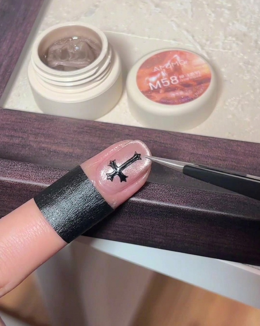 Tweezers placing a small, dark metallic cross charm onto the center of a pink cat eye gel nail.