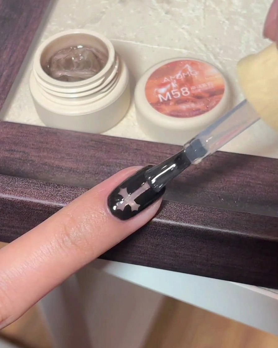 A gel brush applying glossy clear top coat over a black nail featuring a cross charm design.