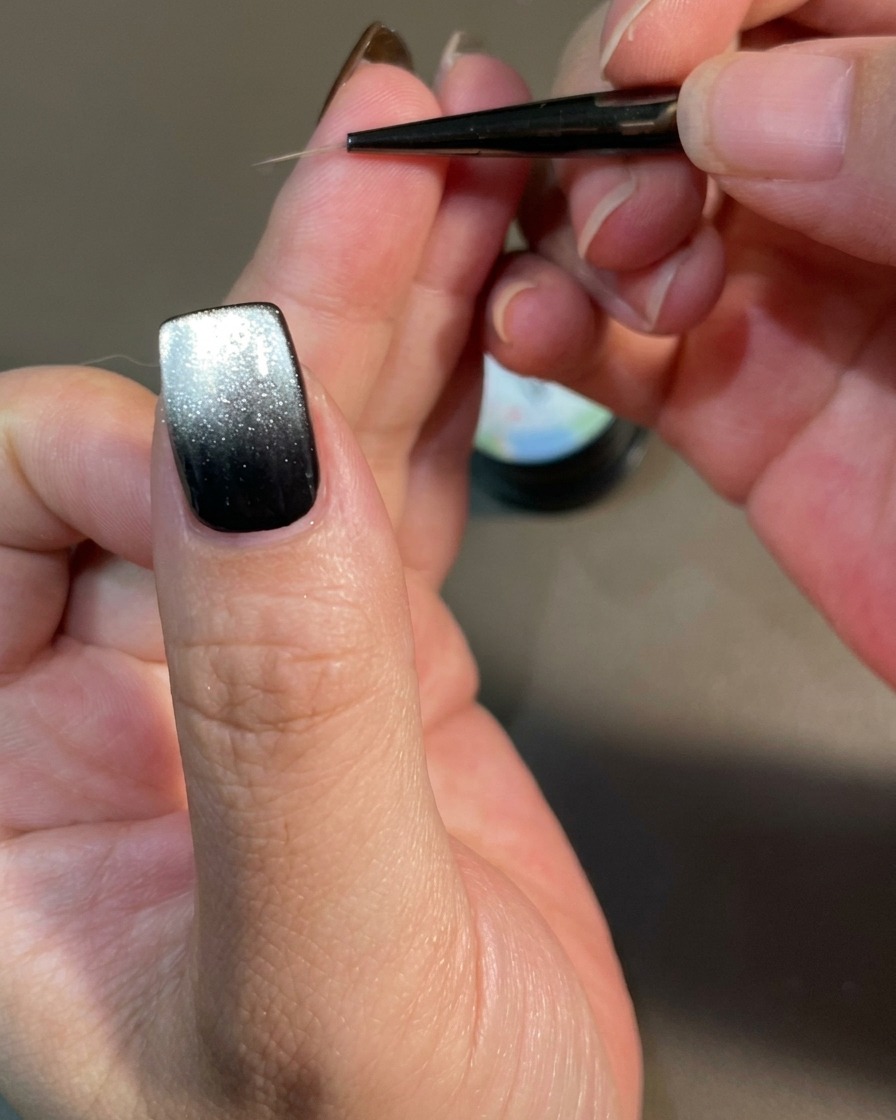 A close-up view of a nail being decorated with a fine liner brush creating curved, flame-like lines on a black magnetic gel base.