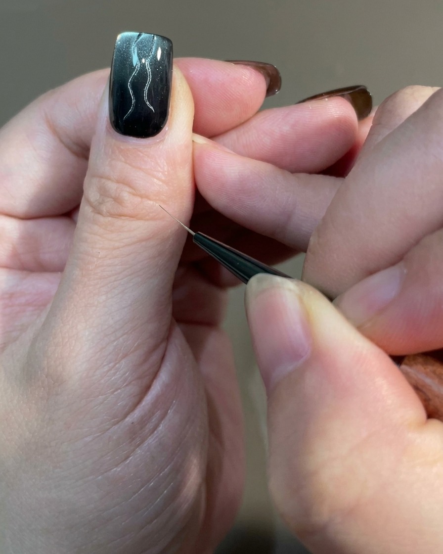 A close-up of a nail showing multiple curved lines of varying lengths being drawn with a liner brush over a magnetic gel base.