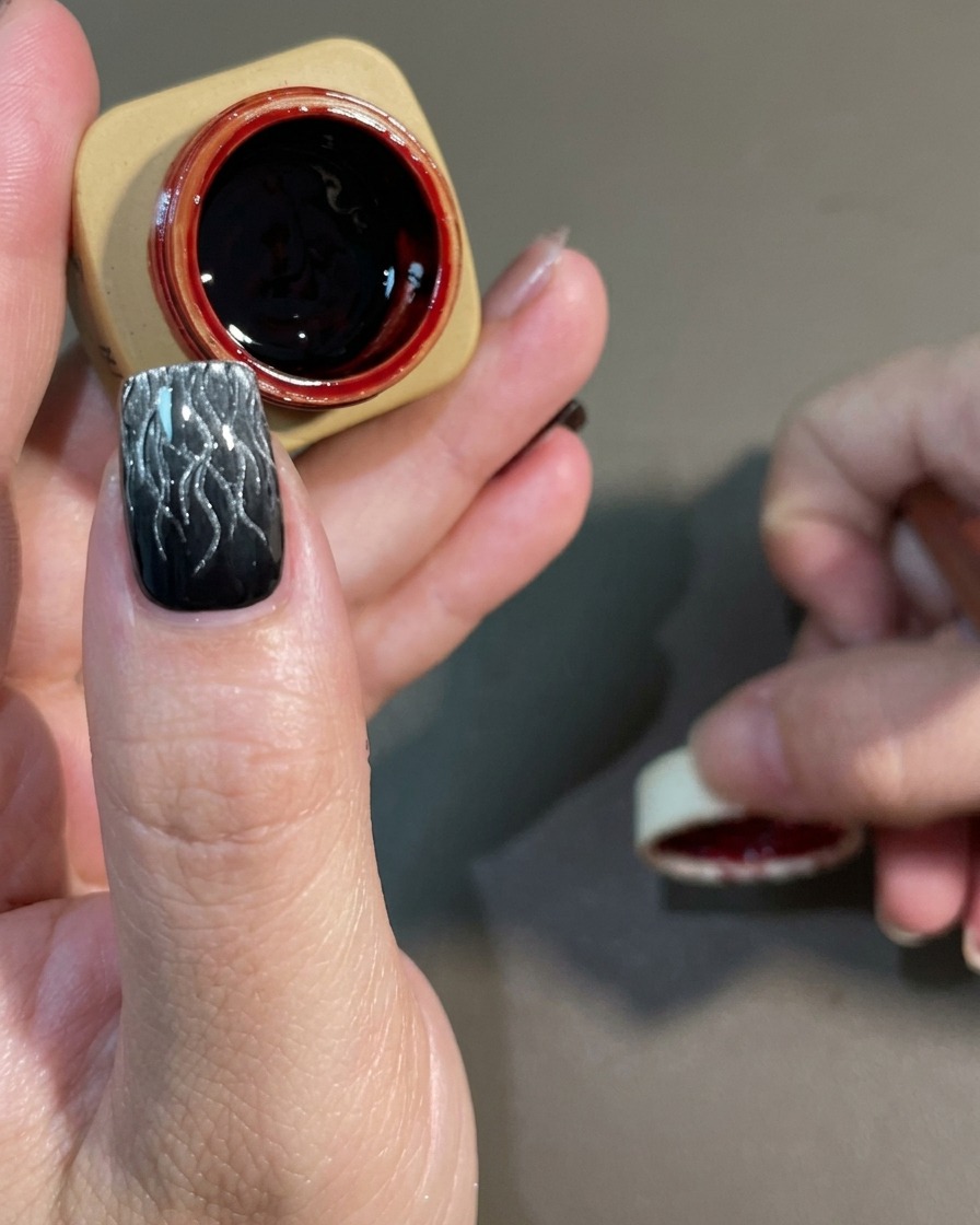 A nail art brush applying a sheer, dark red gel over a cured black and white cat-eye flame design.