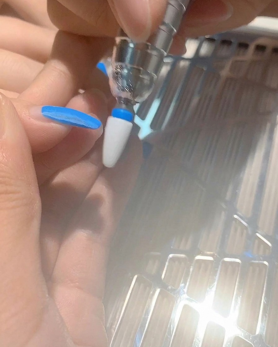 A close-up of a nail drill bit removing blue gel polish from a fingernail.