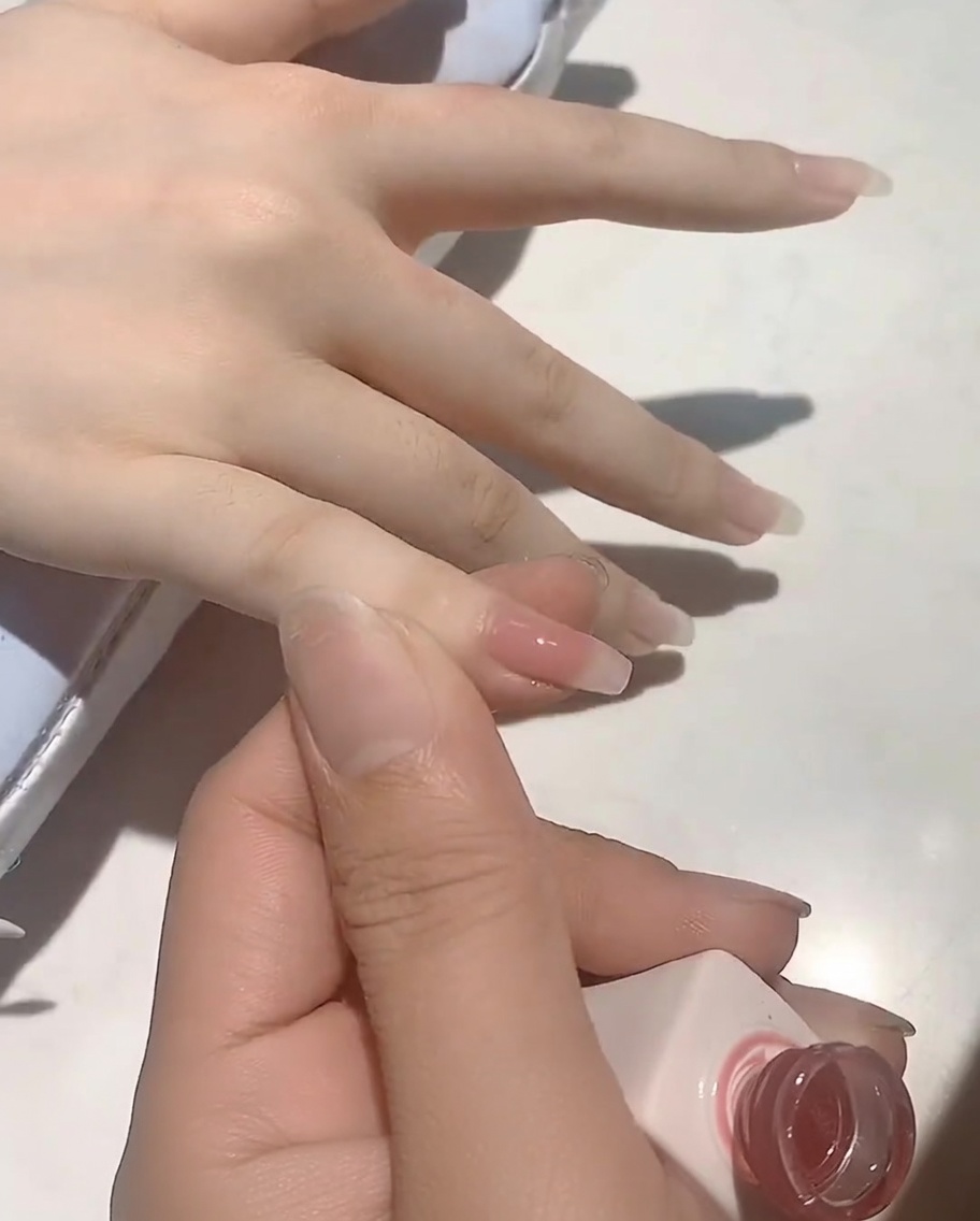 A brush applying a soft nude-pink gel polish to a natural fingernail.