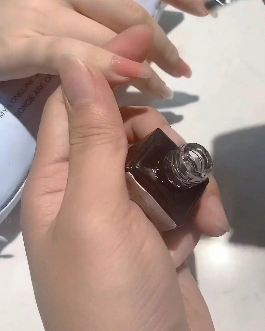 A close-up of a brush applying a clear, glossy top coat to a nude-colored nail.