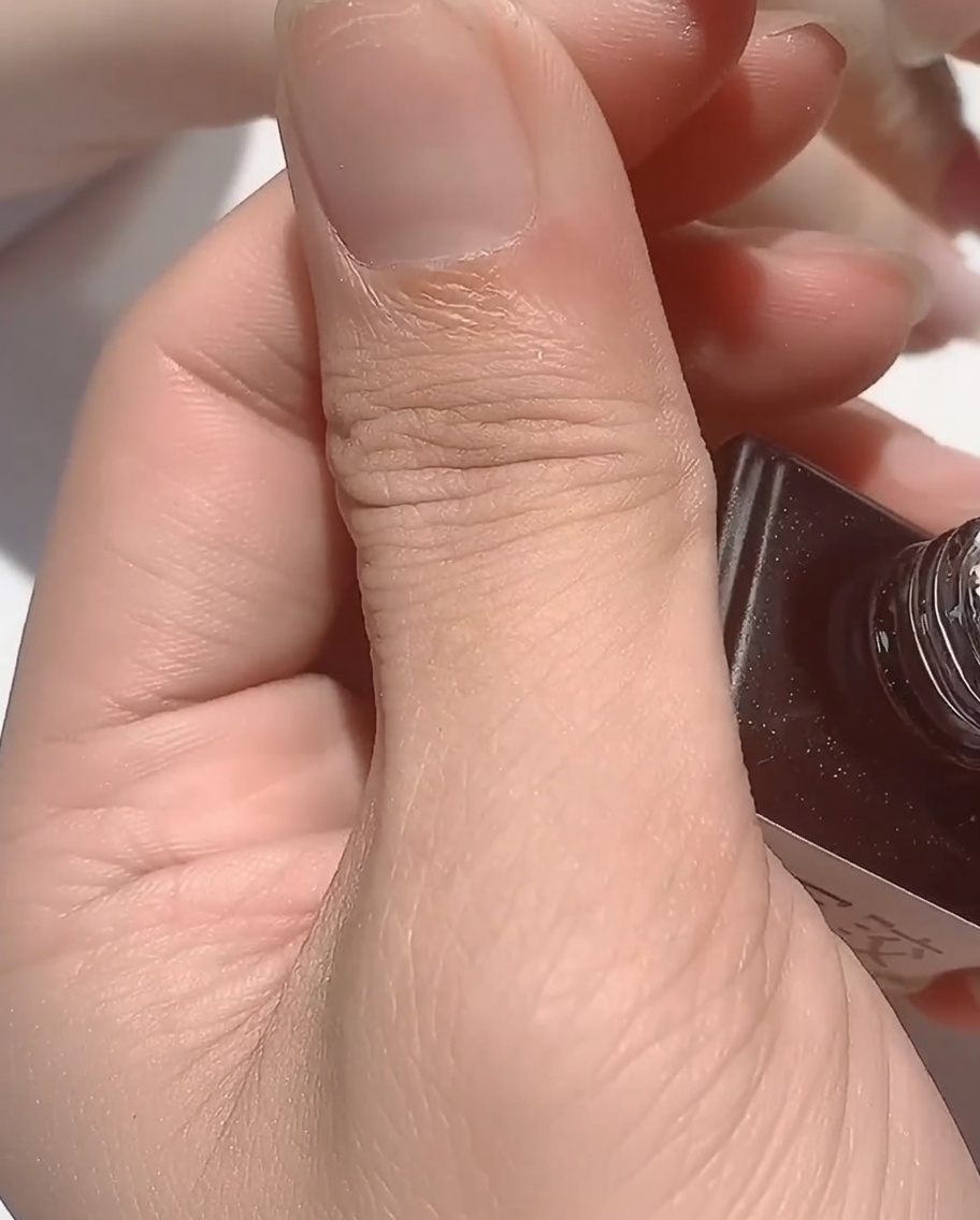 A brush applying a clear, viscous rubber base coat over a nail with iridescent detailing.