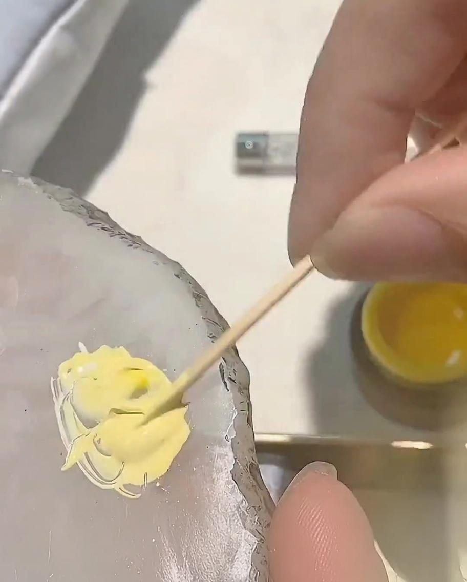 A small dot of red gel polish being added to the existing yellow gel mixture on a palette.