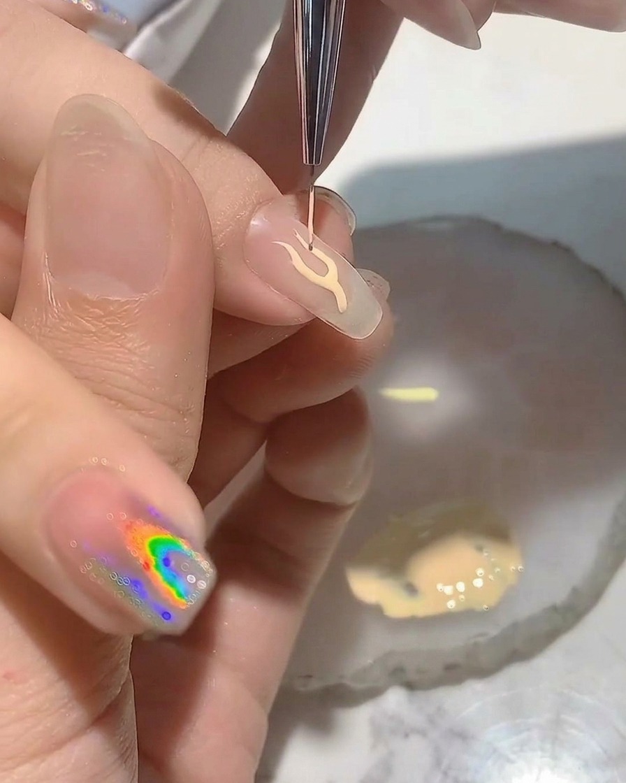 A nail artist using a fine liner brush to paint a delicate flame outline on a clear nail tip.