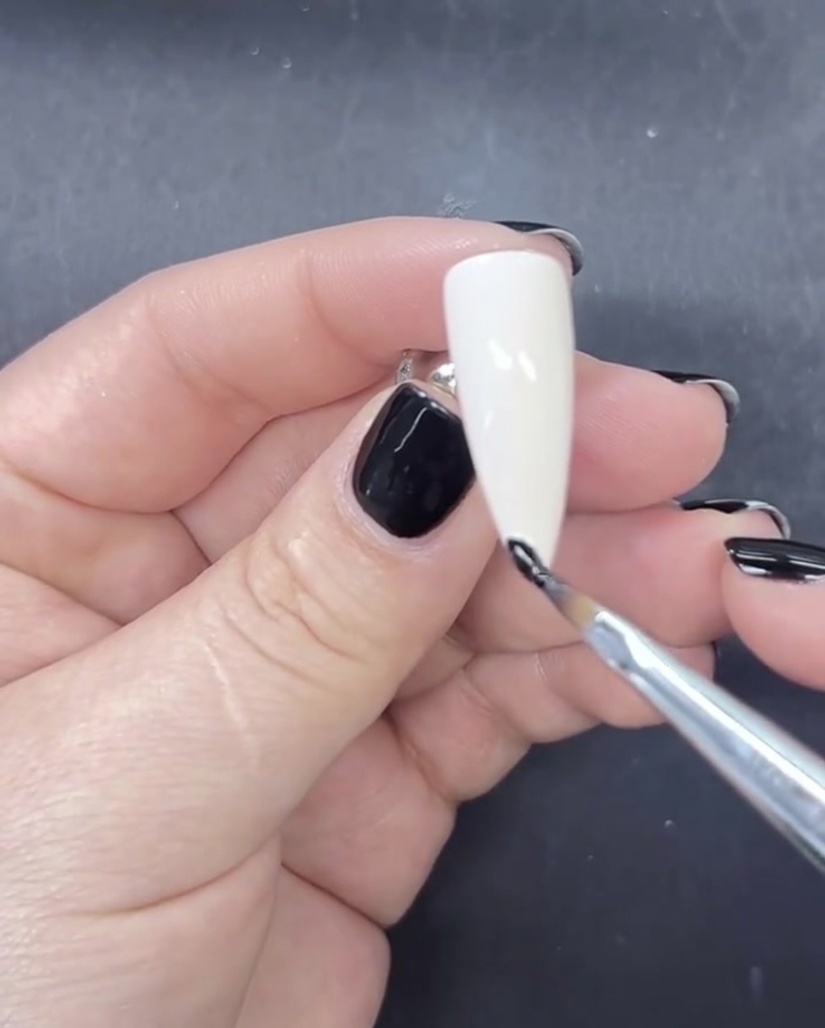 A fine liner brush applying a small, precise line of black gel polish to the tip of a milky white painted nail.