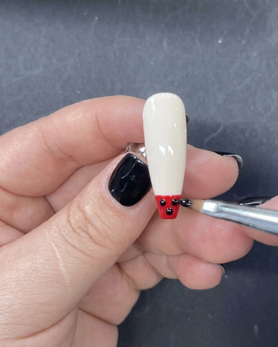 A fine liner brush applying a horizontal strip of red gel polish just above the black tip on the nail.