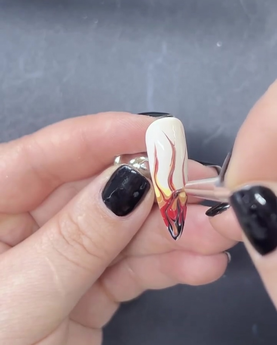 A close-up of a fine liner nail brush pulling red and black gel polish upwards over a white base to create flame shapes.