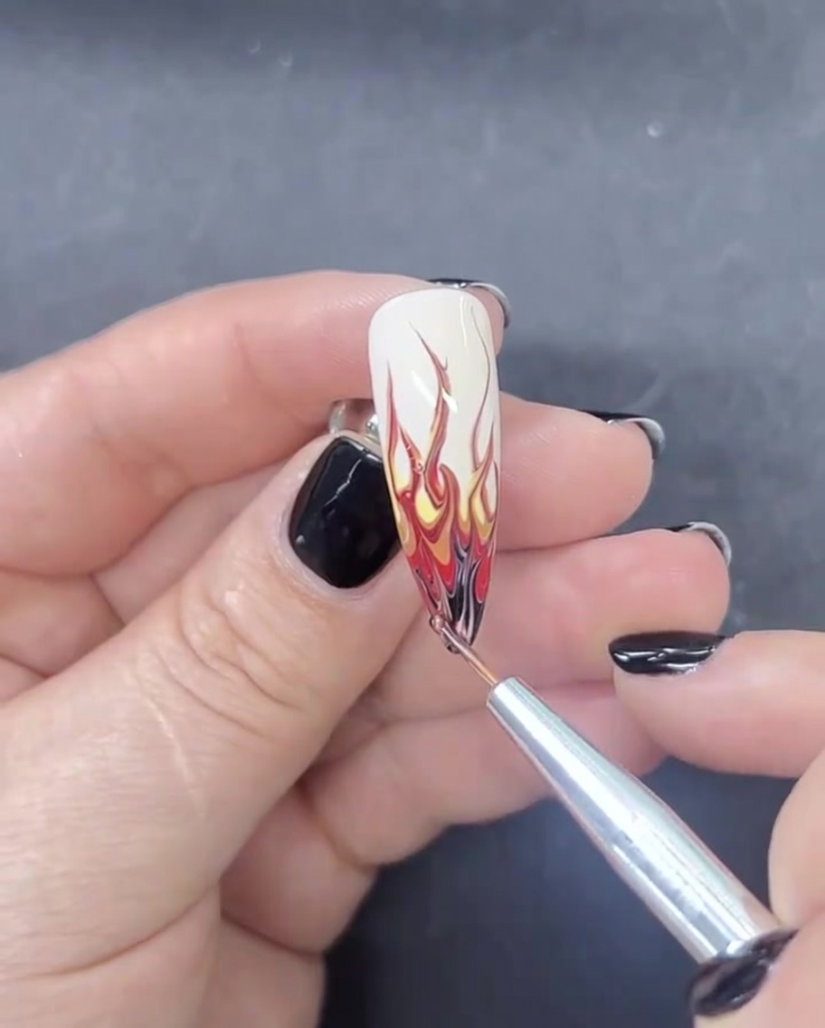 A fine liner nail brush applying a tiny amount of black gel polish to the very tip of an almond-shaped nail to finish a flame design.