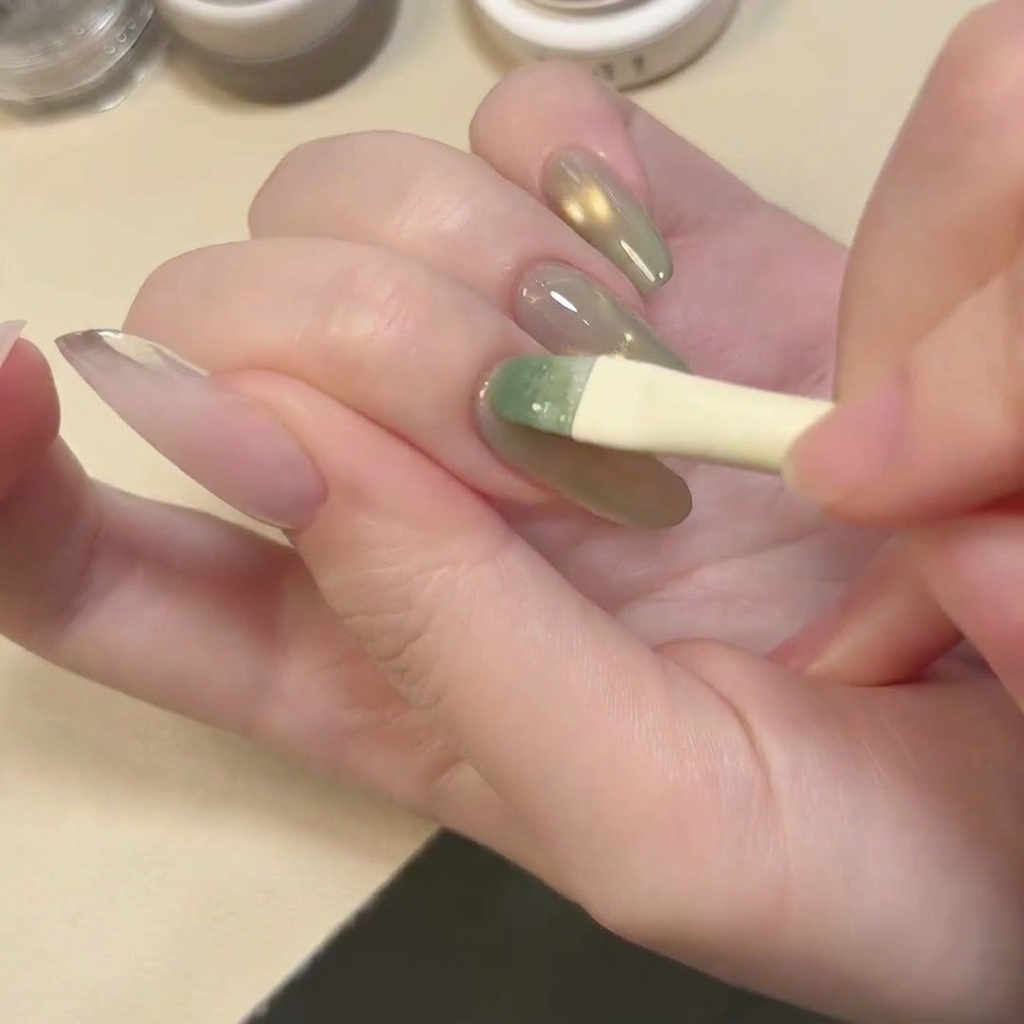 A brush applying a coat of translucent, shimmery lake green gel polish over the nail.