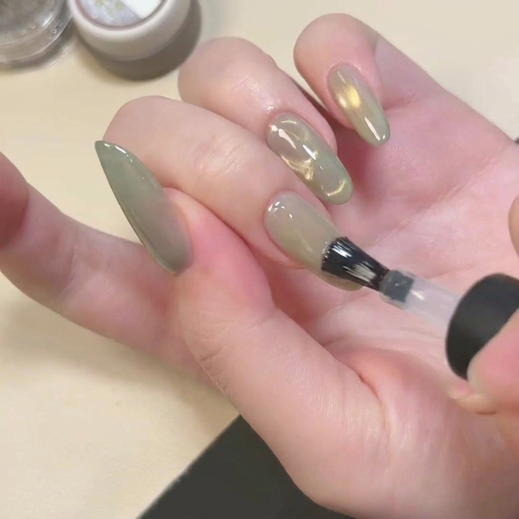 A brush applying a clear, matte-finish top coat over a green cat eye nail.