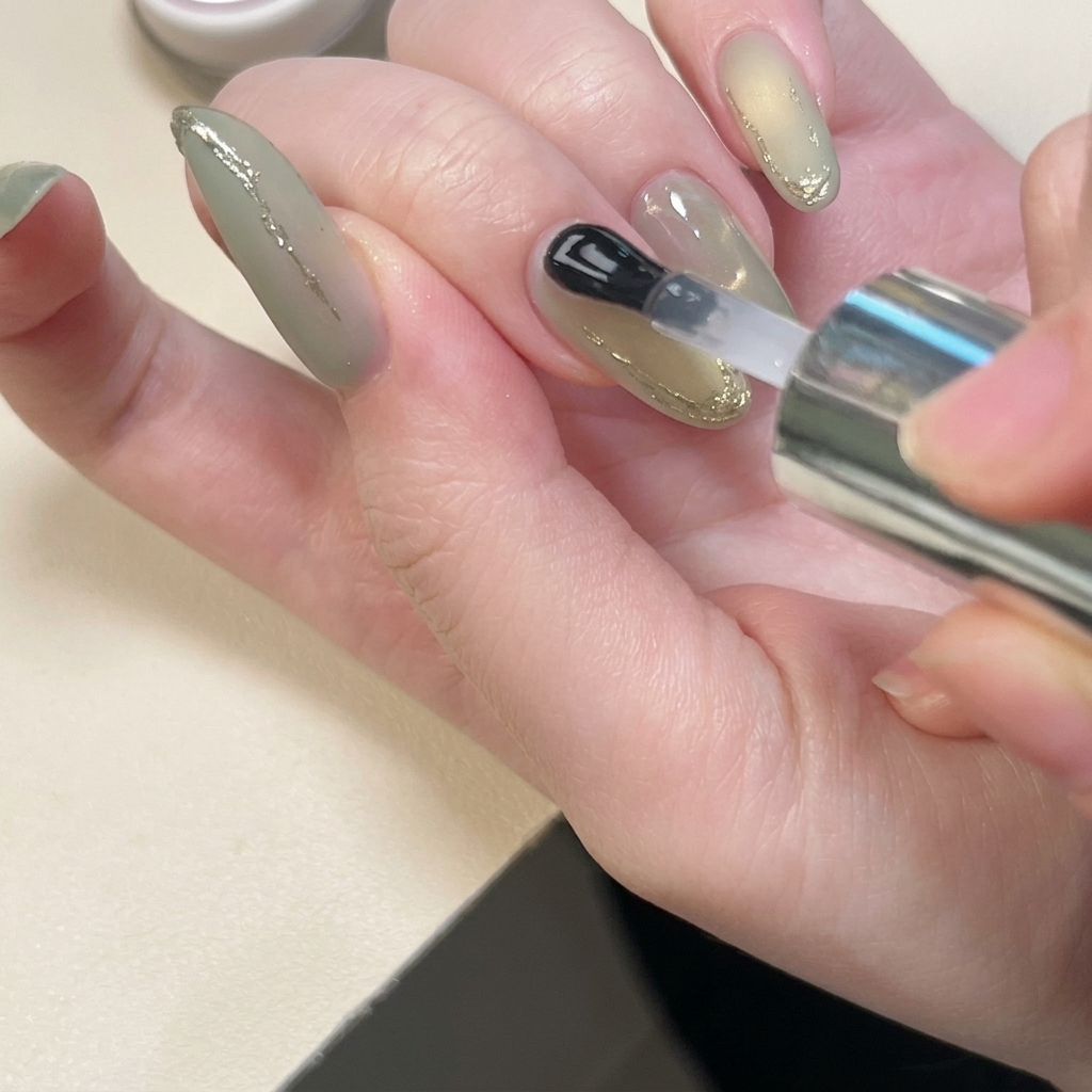 A brush applying a precise layer of clear reinforcing gel over the gold-dusted 3D lines on a fingernail.