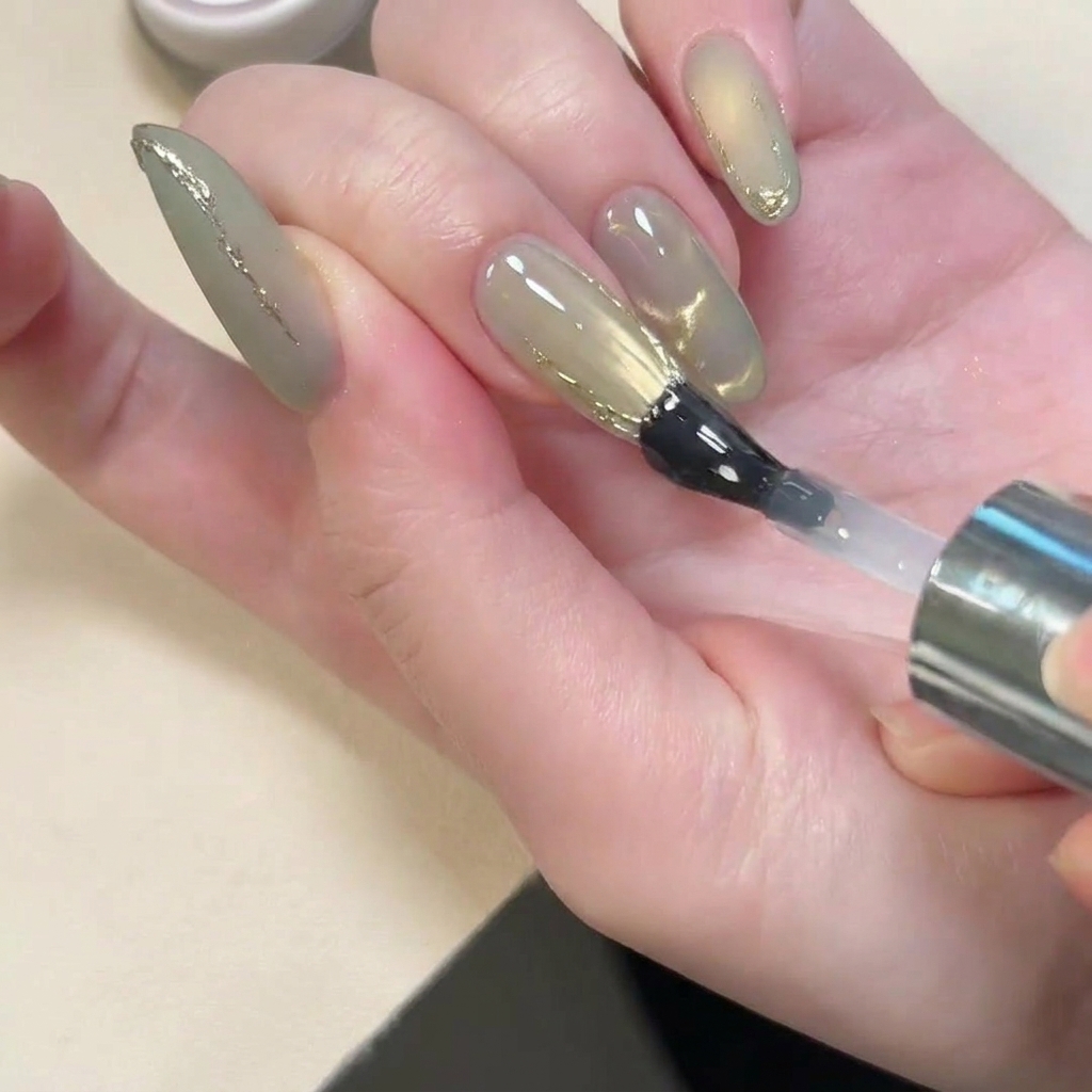 A brush applying a glossy top coat over a nail that has a matte finish and gold 3D accents.