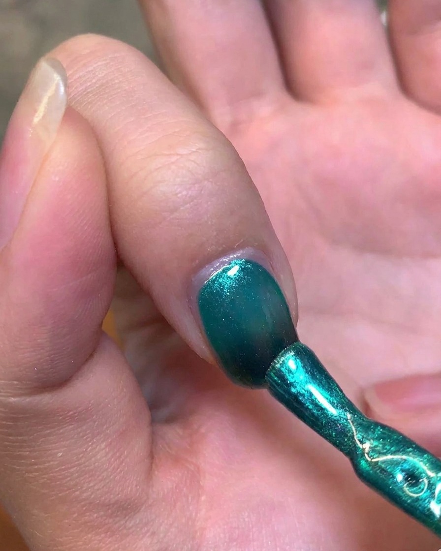 A metal magnetic wand held near the side of a fingernail to manipulate the green gel into a cat-eye highlight.