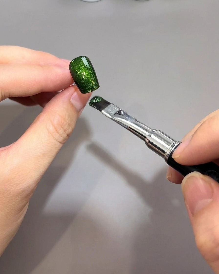 A brush applying a vibrant emerald green magnetic gel over a black nail base.
