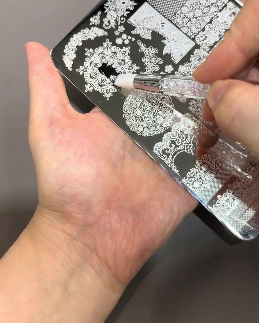 A brush applying thick black polish onto a silver metal nail stamping plate with intricate lace and floral patterns.