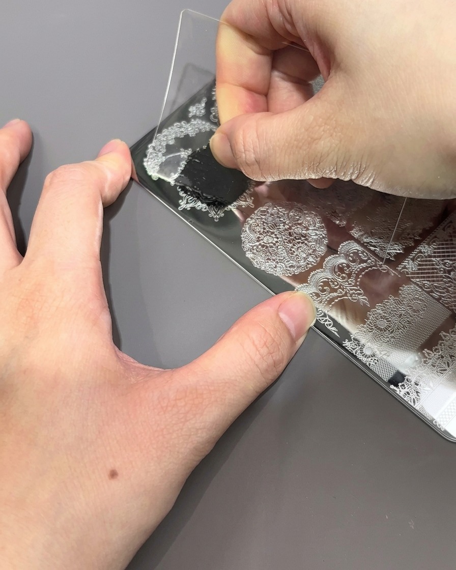 A clear plastic scraper card being pulled across a metal stamping plate, pushing away excess black polish to reveal the engraved pattern.