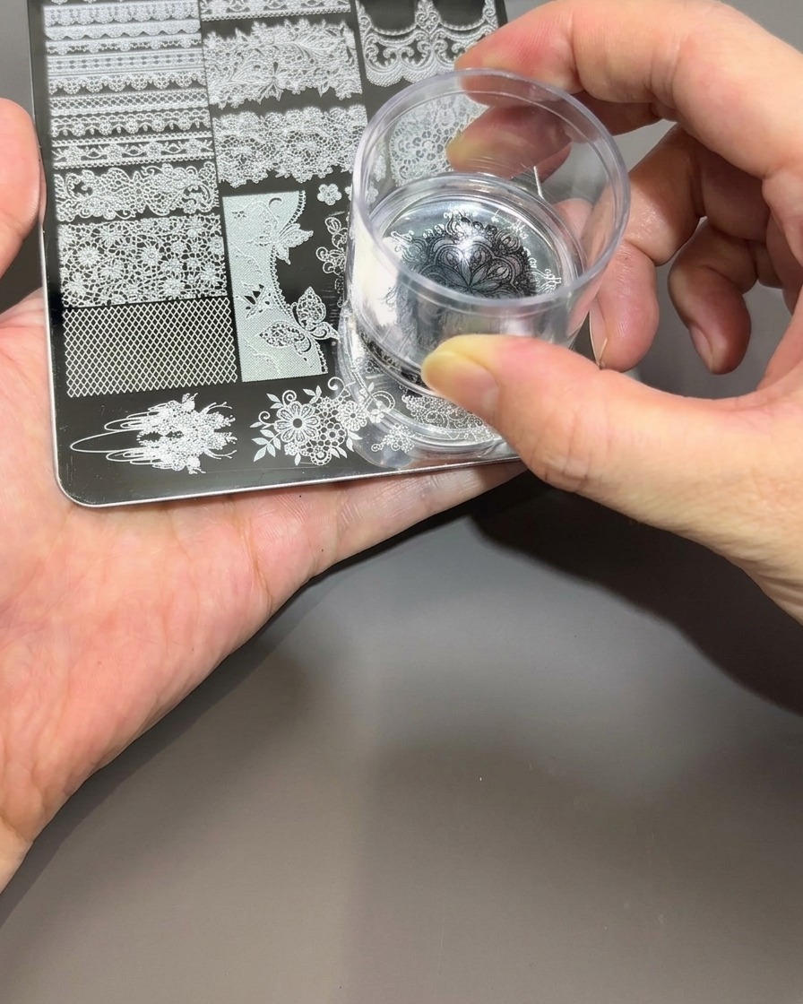 A hand holding a clear silicone nail stamper, gently pressing it onto the black polish design on a metal stamping plate.