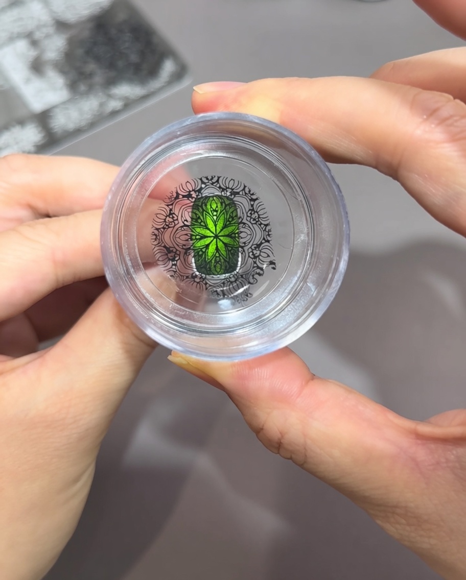 Looking straight down through a clear silicone stamper, showing an intricate black pattern perfectly aligned over a bright green cat-eye nail.