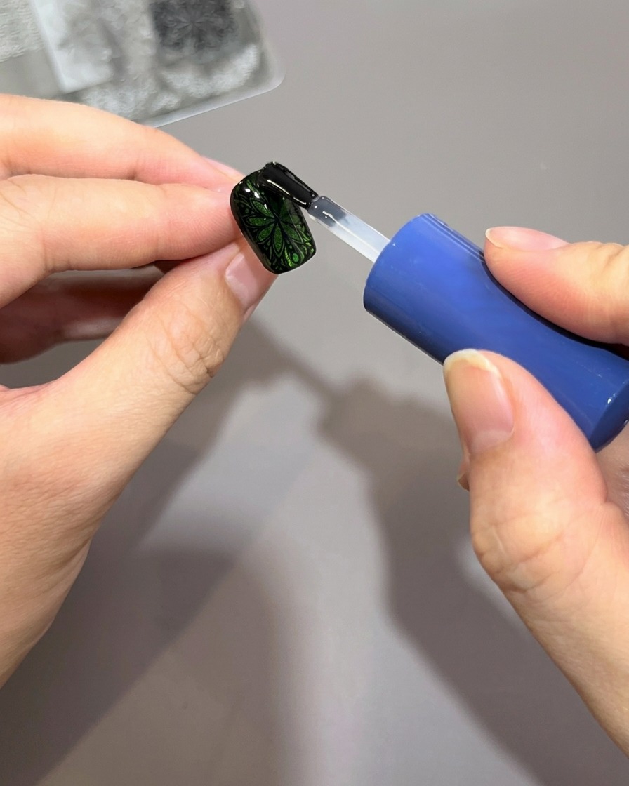 A nail polish brush applying a thick layer of clear glossy top coat over a green nail decorated with a black intricate stamped pattern.