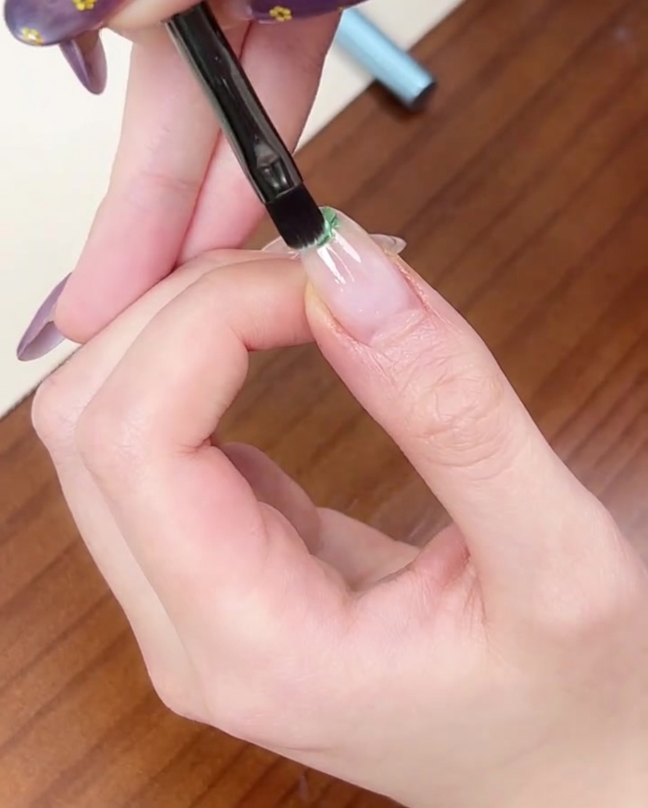 A flat brush applying a small dot of light green gel polish to the tip of a natural nail.