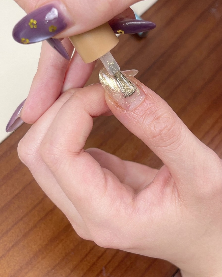 A brush applying a coat of shimmery pink-gold cat-eye gel over the nail.