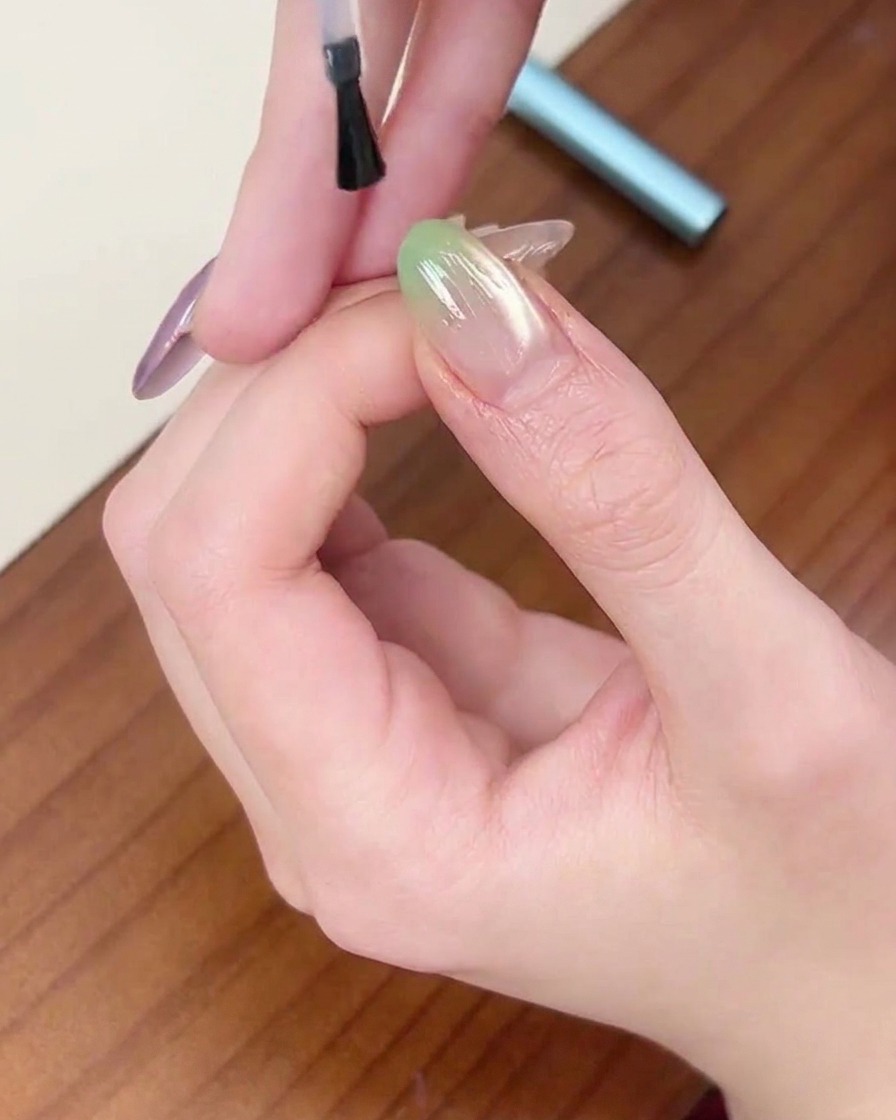 A brush applying a final layer of clear top coat over a green gradient nail.