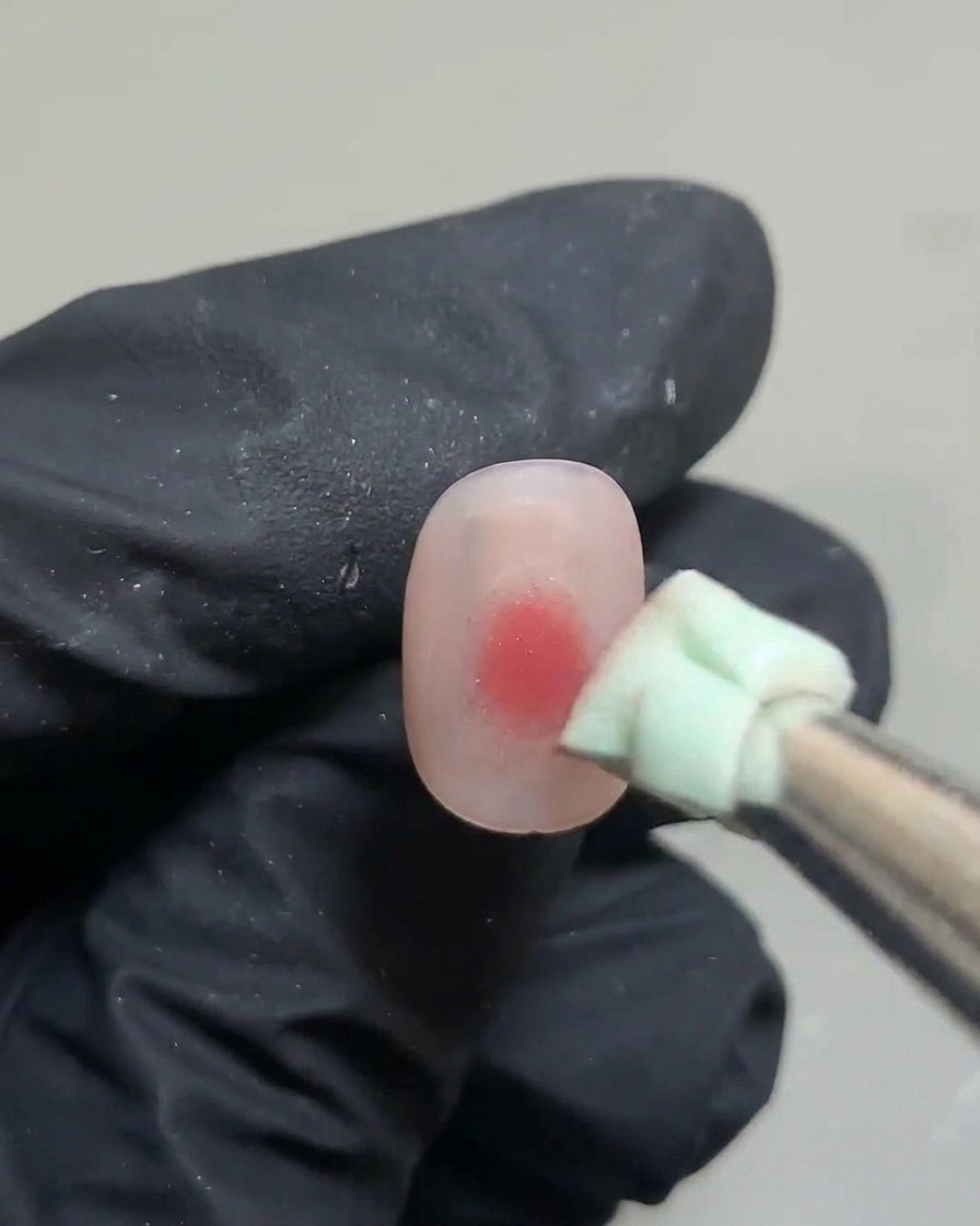 A pair of tweezers holding a small piece of green sponge, patting down a pink gel dot on a nail.