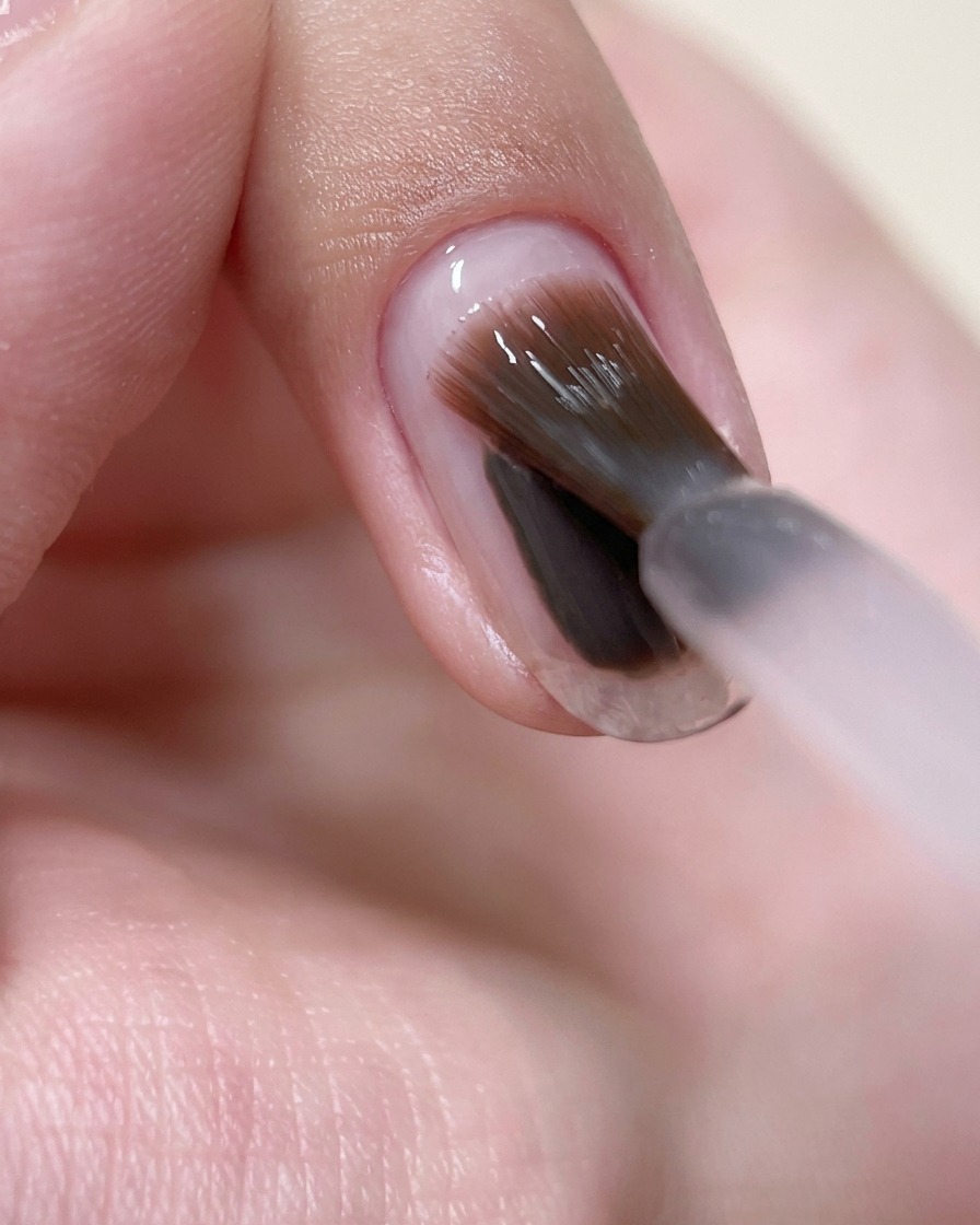 A close-up view showing a brush applying a thin layer of sheer pink builder gel over a natural fingernail.