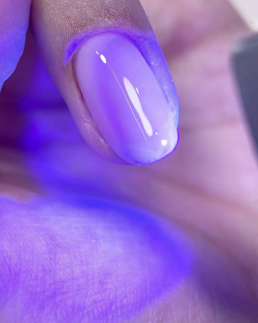 A fingernail with an uncured layer of glossy pink gel resting under the bright purple light of a UV/LED nail curing lamp.