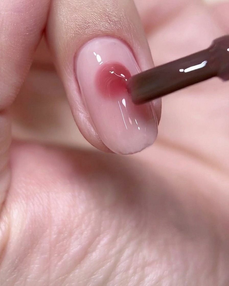 A nail brush depositing a precise, round bead of deep rose-wine colored gel onto the center of a light pink nail base.