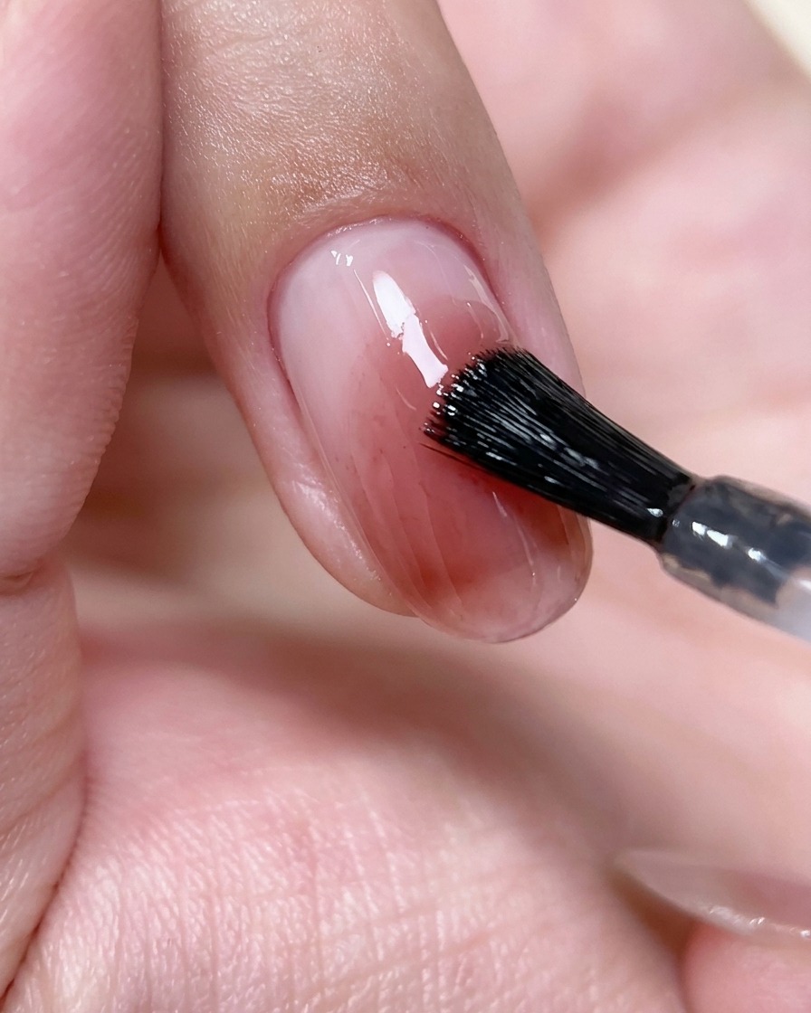 A nail brush applying a thin, clear layer of top coat over a pink blush nail design.