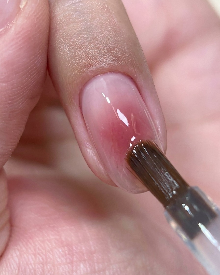 A nail brush depositing a small bead of clear top coat gel onto the center of a glossy pink nail.