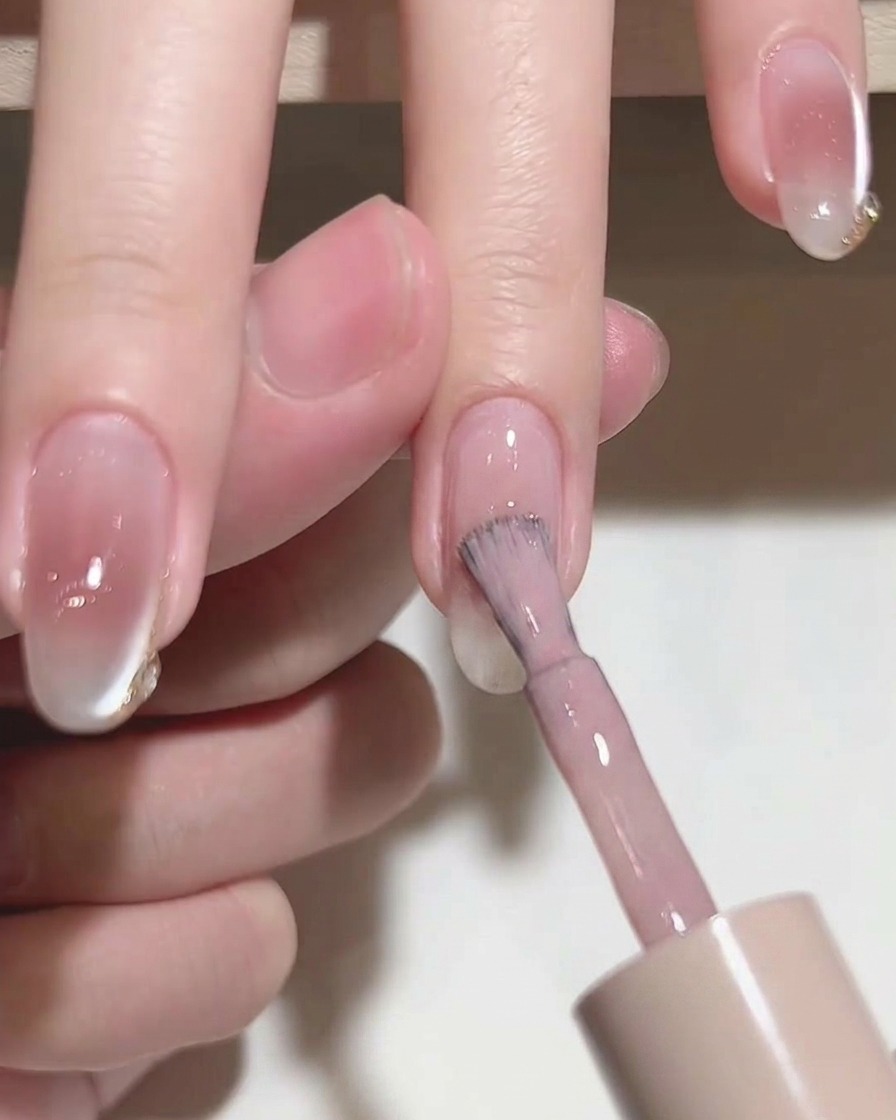Close-up of a brush applying translucent pink gel polish to a natural nail.