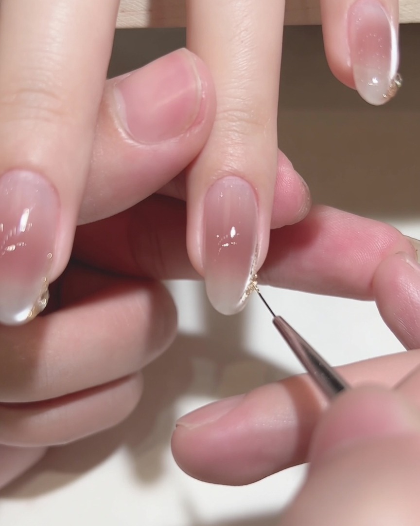 A close-up shot of a fine liner brush applying gold detail to a manicured nail tip.