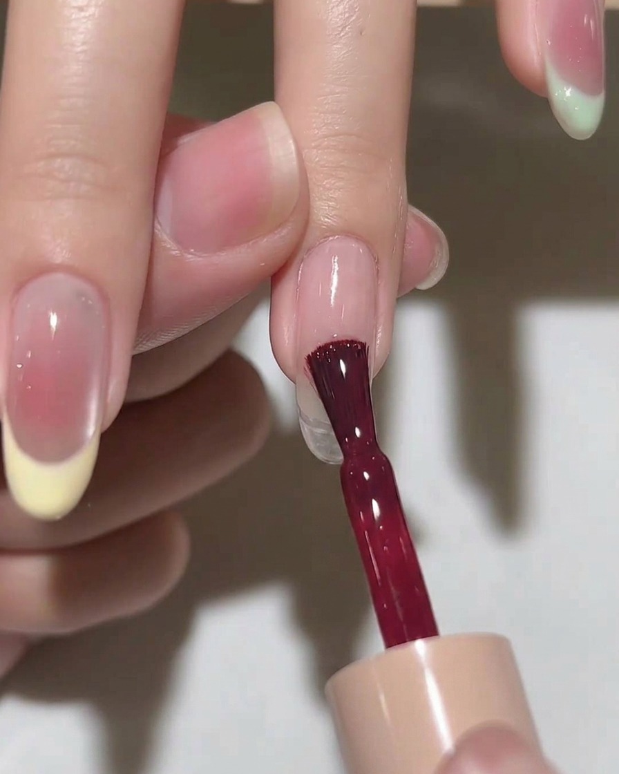 A brush applying a coat of sheer pink gel polish to an almond-shaped nail.