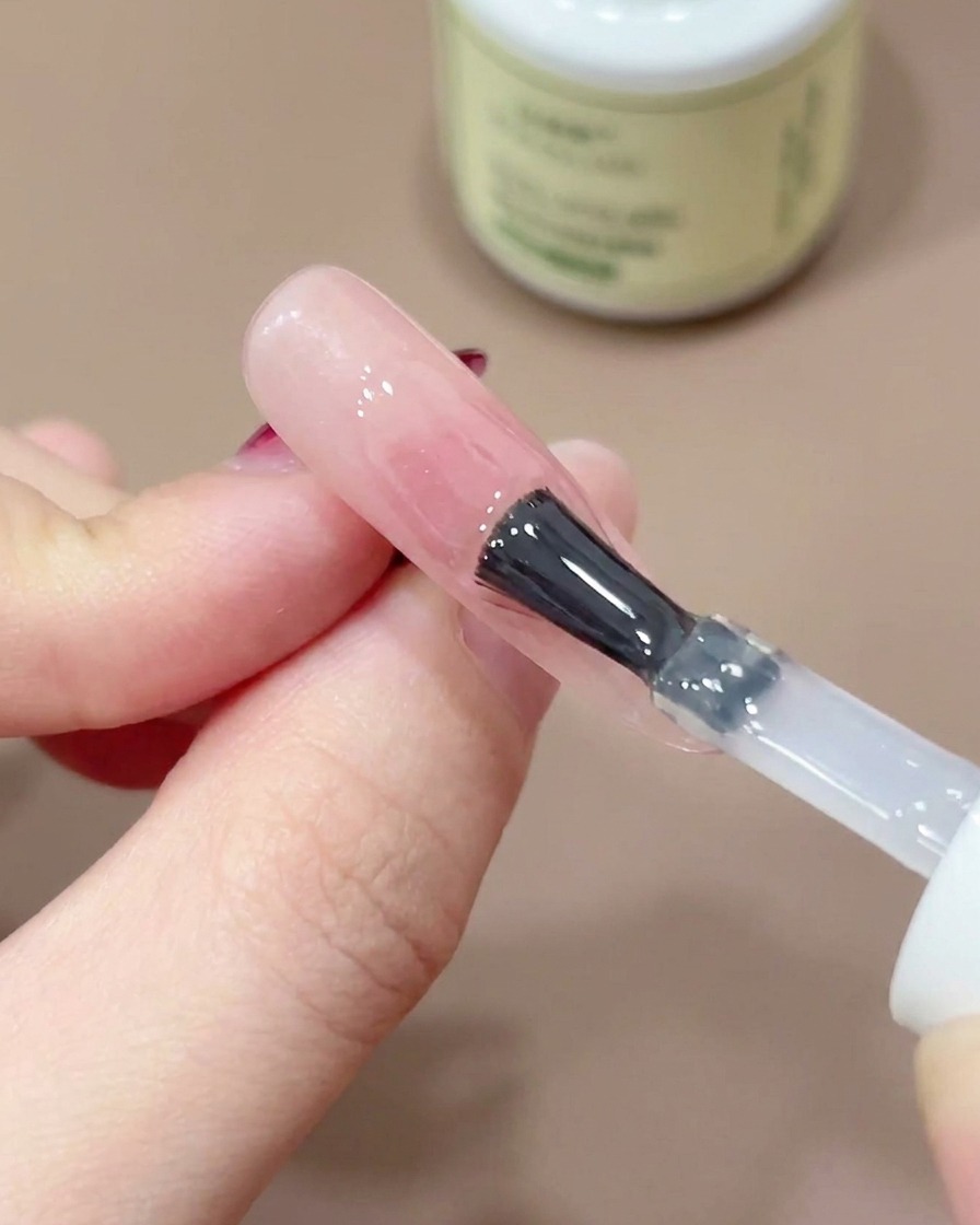 A nail brush applying a clear, glossy top coat over a pink blush nail design.